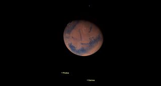 a dark rusty red Mars with dark blue seas hangs in black space. tiny points below are labeled Phobos and Deimos.