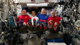 four people in red, white and blue shirts float in zero gravity in a cramped laboratory filled with wires and computers