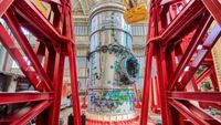 The cylindrical HALO module is seen standing vertically, surrounded by a red steel frame. The HALO module is silver and is lined with coils of green wires. ⁣