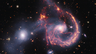 A pair of interacting galaxies. The larger of the two galaxies is slightly right of centre, and is composed of a hazy, bright, white centre and a ring of gaseous filaments, which are different shades of red and orange. Toward the bottom left and bottom right of the ring are filaments of gas spiralling inward toward the core. At the top left of the ring is a noticeable gap, bordered by two large, orange pockets of dust and gas. The smaller galaxy is made of hazy and white gas and dust, which become more diffuse further away from its centre. To this galaxy’s bottom left, there is a smaller, more diffuse gas cloud that wafts outward toward the edges of the image. Many red, orange, and white galaxies are spread throughout, with some being hazier in appearance and others having more defined spiral patterns.